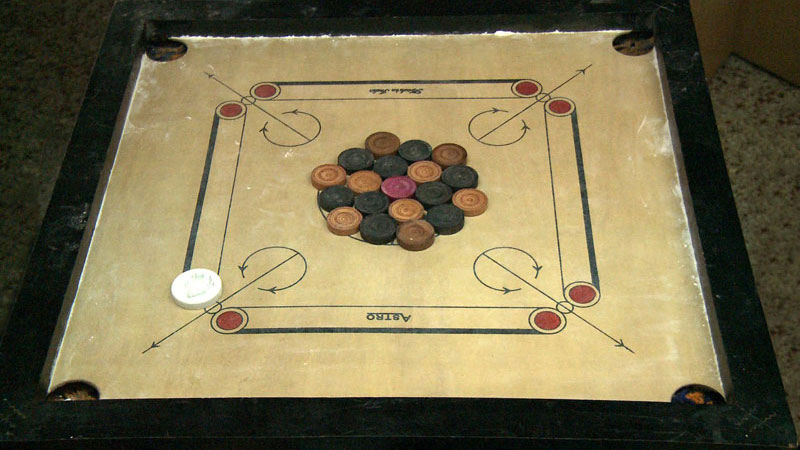 Carrom board What is Carrom board? Carrom board is a board game that is somewhat similar to billiards or snooker but it is not played by balls and sticks. It is very much popular in most of the South East Asian countries like. • Pakistan • Bangladesh • Sri Lanka • Japan • Malaysia • Indonesia. There are carom board cafes in these countries and they regularly organize carom board competitions. Apart from Asian countries, carrom board game gain popularity in Europe and North America too. Countries like Italy, UK, France, Germany, Greece, Spain, Switzerland, etc. also formed their carrom federations. Even Australia and the United States have picked up the like of Carrom and is growing rapidly amongst the citizens. A Complete More About Carrom board Game History In the year 1988, the International Carrom board Federation or ICF was founded in the city of Chennai in India. The formal rules for the game were published in the same year. After formal rules were established the Indian Diaspora spread the game in Europe and in the US and Canada also. How to Play Carrom board? The carom board is played on plastic or (mostly) wooden carrom boards of different sizes. The objective is to push the lightweight wooden discs which are called the ‘carrom men’ into the four different pockets on the four corners of the carrom board with the help of a striker, which is made of plastic. The carrom men are in three different colors, Black, White, and Pink. Total 9 black and 9 white are fixed in such a way that it makes a circle in the middle of the board and in the very center, the pink carrom man called the Queen is put. As per ICF-approved measurement, the Carrom men should between 3.02 – 3.18 cm in diameter. The Queen can be won only if it is covered by another carrom man in consecutive shots. To make the carrom board surface smooth, boric acid powder is often used in India. Types of Carrom board Family Point Carrom board The game of Carrom board originated as a family game, hence the first variant is obviously related to how people play the game of Carrom board at their homes. The main objective of this variant is to enjoy and spend some quality time with the family. Here there is no stoppage on which color disks you need to pocket, for each black disk pocketed the player gets 5 points, 10 for the white, and queen earns 25 points for the player provided if it is covered. The highest number of point winners ultimately wins the carom board game. This game can be played individually or also in pairs as well. Point Carrom board This is quite similar to the family point carom board but over here both the black and white disks will earn you 1 point each, whereas after covering the queen it will earn you 3 points. The player who reaches the 25 points mark is declared the winner and if there is no clear winner then the highest number of point winners will be declared as the winner. In case of a tie, a tiebreaker is played where players are allowed to choose two different numbers of disks and they need to pocket them only on rebounds. Total Point Carrom board In this variant which is very much popular in India and Pakistan, two players play individually and turn by turn. The black disks get you 5 points and whites will worth 10 points. 50 points will be earned if the queen has been properly covered. The players keep on playing carrom board till all the Carrom men are pocketed by the lowest scorer player after the first round. Professional Carrom board These carrom board rules are formed by the ICF and for the national and international Carrom championships, these carrom rules are used. As per these rules, every Carrom game consists of 29 points. Each player is assigned with a color of disks and they have to pocket the assigned color disks only. The players can’t pocket the queen before the first disk is pocketed and after the last disk is pocketed. As per normal rules over here too the queen must be duly covered. The queen and its cover can’t be pocketed in the same pocket. The first person who pockets all of his designated color disks is declared the winner.